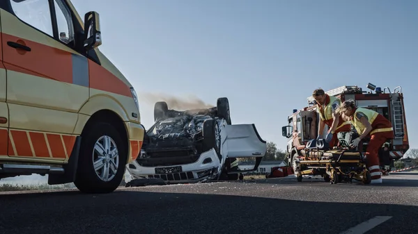 Equipe de resgate de bombeiros e paramédicos Trabalhar em uma cena terrível acidente de carro. Preparando equipamentos, alongamentos, primeiros socorros. Salvando pessoas feridas e presas do veículo em chamas — Fotografia de Stock