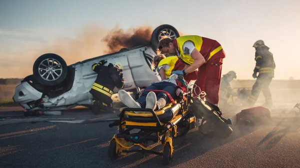 On the Car Crash Traffic Accident Scene: Paramedici redden het leven van een vrouwelijk slachtoffer liggend op Stretchers. Ze luisteren naar een hartslag, brengen zuurstofmasker aan en geven EHBO. Brandweerlieden blussen vuur — Stockfoto