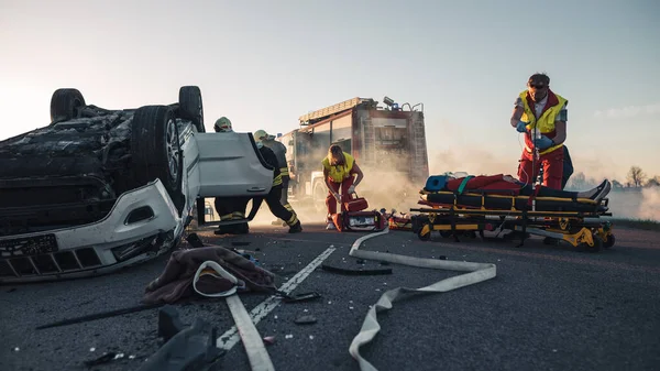 Paramedici en brandweerlieden arriveren op de auto-ongeluk plaats delict. Professionals redden verwond slachtoffer gevangen in roll-over voertuig door het bevrijden van hen, het geven van EHBO en blussen van brand — Stockfoto