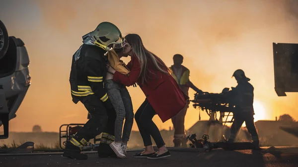 Dappere brandweerman draagt gewond jong meisje naar veiligheid waar ze herenigd met haar liefhebbende moeder. Op de achtergrond Auto crash Verkeersongeval Courageous Paramedici en brandweerlieden redden levens — Stockfoto