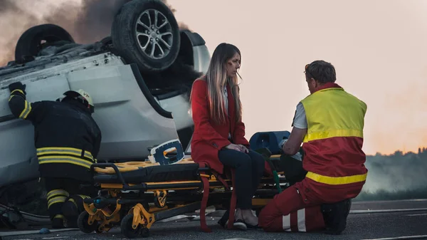 På bilolyckan Trafikolycka Scen: Paramedicin Utför första hjälpen av en kvinnlig offer som sitter på bårar. Rulla över i bakgrunden. — Stockfoto