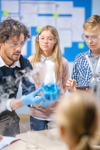 Grundskola Science Classroom: Entusiastisk lärare förklarar kemi för olika grupper av barn, visar dem hur man blandar kemikalier i bägare. Barn använder digitala surfplattor och pratar — Stockfoto