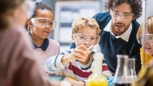 Aula de Ciencias de la Escuela Primaria: Profesor Entusiasta Explica Química a Diverso Grupo de Niños, Niño Pequeño Mezcla Químicos en Beakers. Niños aprenden con interés —  Fotos de Stock