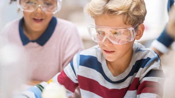 Портрет мальчика-энтузиаста в защитных очках смешивает химикаты в стаканах. Класс естественных наук начальной школы: дети учатся с интересом. — стоковое фото