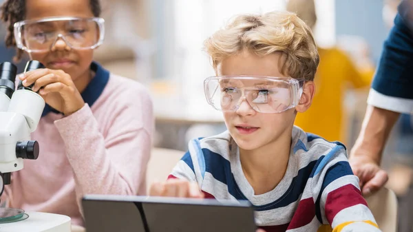 Aula de Ciencias de la Escuela Primaria: Boy utiliza Tablet Computer digital para comprobar la información en Internet mientras que Entusiasta linda niña utiliza microscopio. —  Fotos de Stock