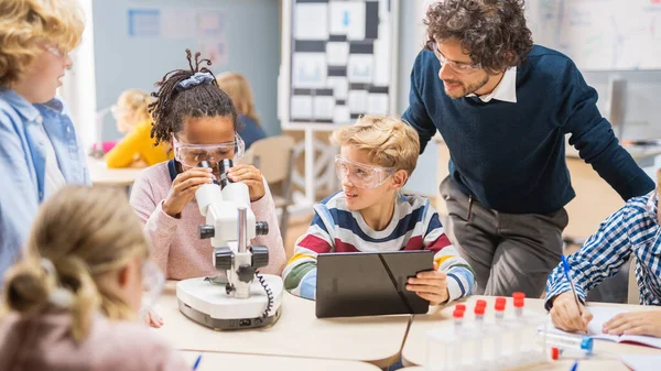 Elementary School Science Classroom: Söt liten flicka ser ut under mikroskop, pojke använder digitala surfplatta dator för att kontrollera information på Internet, medan Entusiastiska läraren förklarar Lesson — Stockfoto