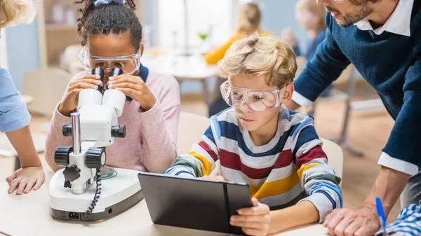 小学科学课室：可爱的小女孩在显微镜下看，男孩使用数字平板电脑在网上查看信息，而热心的教师则帮助男孩 — 图库照片