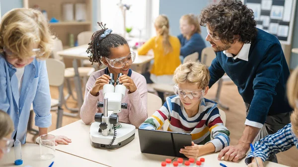 小学科学课室：可爱的小女孩在显微镜下看，男孩使用数字制表计算机在网上查看信息。老师从后面看 — 图库照片