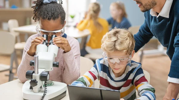 Aula de Ciencias de la Escuela Primaria: Linda Niña Mira Bajo Microscopio, Boy Utiliza Tablet Computadora Digital para Comprobar la Información en Internet. Profesor observa desde atrás — Foto de Stock