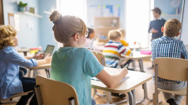 Clase de Ciencias de la Escuela Primaria: Linda Niña está escuchando a un maestro y tomando notas. Profesor de Física Explica Lección a una Clase Diversa llena de Niños Inteligentes. Sobre el hombro disparado. —  Fotos de Stock