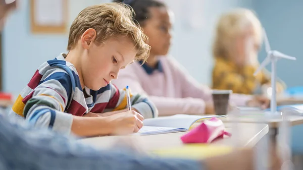 Dalam Elementary School Classroom Brilliant Caucasian Boy Writes in Exercise Notebook, Taking Test and Writing Exam. Kelas Junior dengan Kelompok Anak-anak Bekerja Diligen dan Belajar Barang Baru — Stok Foto