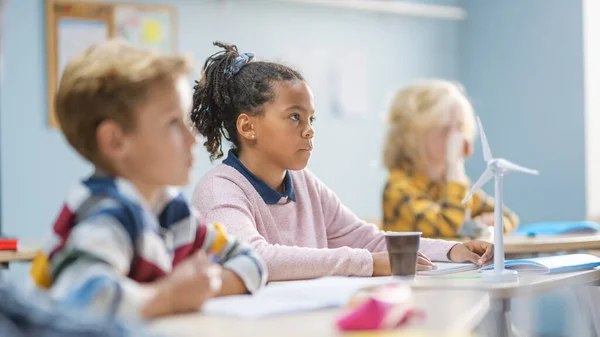 В начальной школе Блестящая чёрная девочка внимательно слушает учителя. Младший класс с группой ярких детей, работающих сообща и создающих новые вещи — стоковое фото