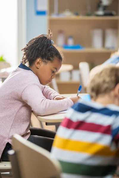 In Elementary School Classroom Brilliant Black Girl Writes in Exercise Notebook, Taking Test and Writing Exam. Junior Classroom with Group of Bright Children Working Diligently and Learning New Stuff — Stock Photo, Image