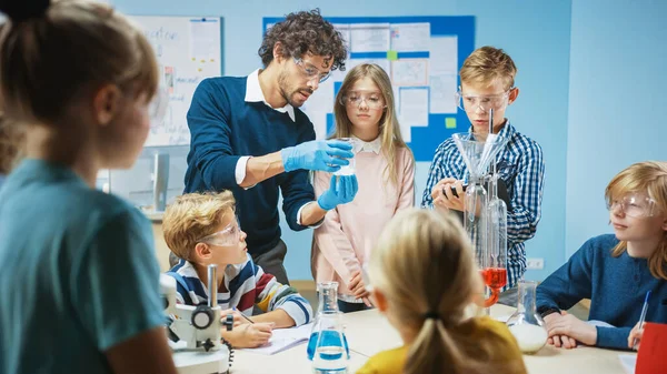 Elementary School Science Classroom: Enthusiastic Teacher explica la química a diversos grupos de niños, les muestra cómo mezclar productos químicos en vasos de precipitados. Niños usan gafas de seguridad y hablan —  Fotos de Stock