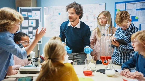 Basisschool Science Classroom: Enthousiaste leraar legt scheikunde uit aan diverse groep kinderen, toont hen hoe ze chemische stoffen in bekers te mengen. Kinderen gebruiken digitale tabletcomputers en praten — Stockfoto