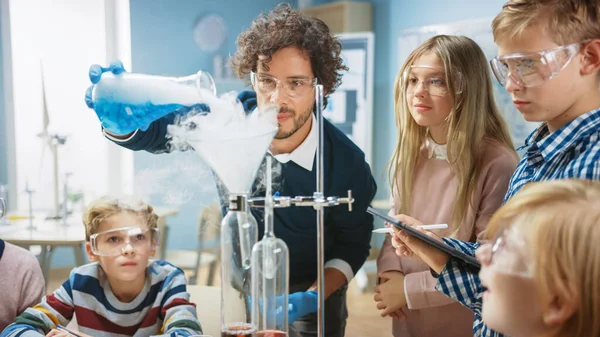 Grundskola Science Classroom: Entusiastisk lärare förklarar kemi för olika grupper av barn, visar dem hur man blandar kemikalier i bägare. Barn använder digitala surfplattor och pratar — Stockfoto