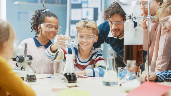Szkoła podstawowa Nauka Klasa: Mały chłopiec miesza chemikalia w zlewkach. Entuzjastyczny Nauczyciel Wyjaśnia Chemię Zróżnicowanej Grupie Dzieci. Dzieci uczą się z zainteresowaniem — Zdjęcie stockowe