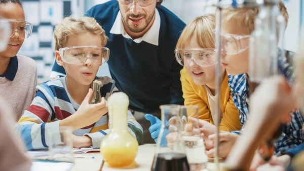 Aula de Ciencias de la Escuela Primaria: Profesor Entusiasta Explica Química a Diverso Grupo de Niños, Niño Pequeño Mezcla Químicos en Beakers. Niños Conocimiento Acqaering —  Fotos de Stock