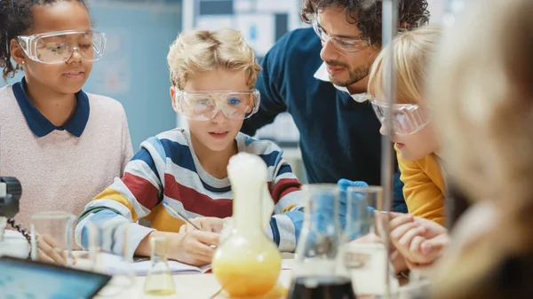 Aula de Ciencias de la Escuela Primaria: Profesor Entusiasta Explica Química a Diverso Grupo de Niños, Niño Pequeño Mezcla Químicos en Beakers, Escribe Resultados. Niños aprenden con interés —  Fotos de Stock