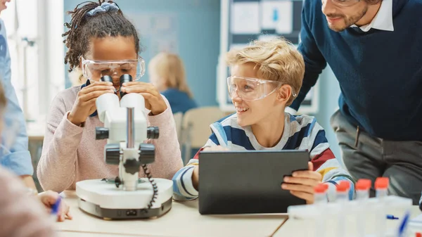 Basisschool Science Classroom: Schattig Meisje kijkt onder de microscoop, Jongen maakt gebruik van Digital Tablet Computer om informatie te controleren op het internet, terwijl Enthousiaste leraar verklaart Les — Stockfoto