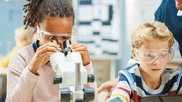 小学科学课室：可爱的小女孩在显微镜下看，男孩使用数字制表计算机在网上查看信息。老师从后面看 — 图库照片