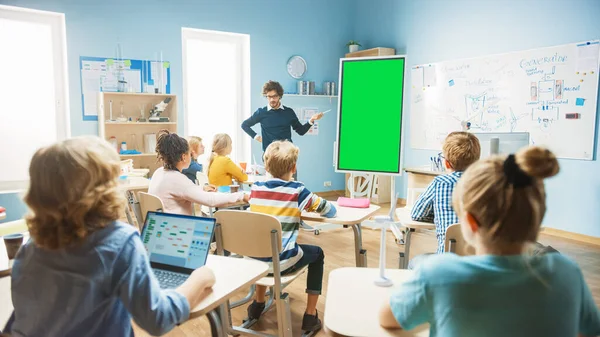小学校物理の先生は緑の画面のモックアップテンプレートとインタラクティブなデジタルホワイトボードを使用しています。彼はスマート多様な子供たちでいっぱいの教室にレッスンをリードしています。子供たちが聞く理科の授業 — ストック写真