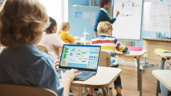 Elementary School Science Class: Over the Shoulder Little Boy använder laptop med skärm Visar programmering programvara. Fysikläraren förklarar för en mångsidig klass full av smarta barn — Stockfoto