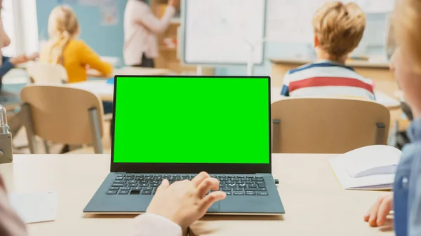 Classe de Ciência da Escola Primária: Pequena menina usar laptop com tela verde modelo Mock-up. Na aula de fundo da escola cheia de várias crianças inteligentes — Fotografia de Stock