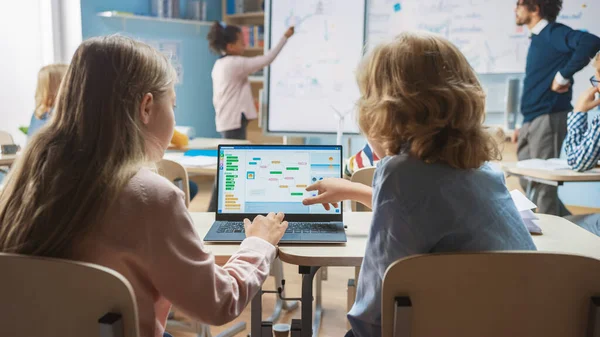 小学校理科の授業:肩の上に小さな男の子と女の子は、画面表示プログラミングソフトウェアとラップトップを使用します。物理学の先生は、スマートキッズがいっぱいの多様なクラスにレッスンを説明します — ストック写真