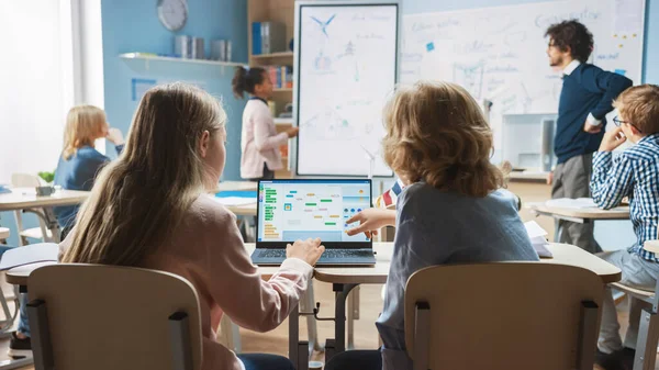 Classe de science de l'école primaire : sur l'épaule, les petits garçons et les filles utilisent un ordinateur portable avec un logiciel de programmation à écran. Un professeur de physique explique la leçon à une classe diversifiée remplie d'enfants intelligents — Photo