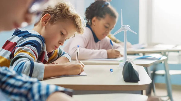 W klasie podstawowej Brilliant Caucasian Boy pisze w zeszycie z ćwiczeniami, zdając test i egzamin pisemny. Klasa młodzieży z grupą dzieci pracujących pilnie i uczących się nowych rzeczy — Zdjęcie stockowe