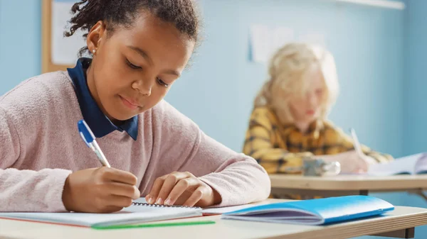 Im Klassenzimmer der Grundschule schreibt ein brillantes schwarzes Mädchen in einem Aufgabenheft, macht Tests und schreibt Prüfungen. Juniorklasse mit einer Gruppe aufgeweckter Kinder, die fleißig arbeiten und Neues lernen — Stockfoto