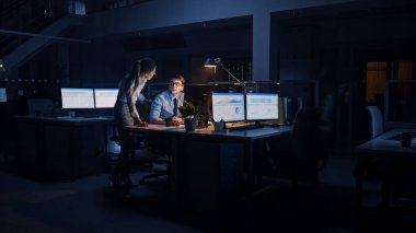 Hırslı ve kendinden emin işadamı Masaüstü Bilgisayarı Kullanıyor, Onun Kadın Proje Yöneticisi Özel Görevleri, Hesaplama ve Stratejik Hareketleri Açıklıyor. Profesyonel İnsanlar Büyük Ofiste Gece Geç Saatlere Kadar Çalışıyor