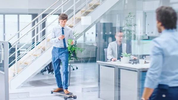 In the Bright Corporate Office Cool Businessman Wearing Tie and Shirt Rides Skateboard through Office Hallway. In the Background Professional People Working on Desktop Computers