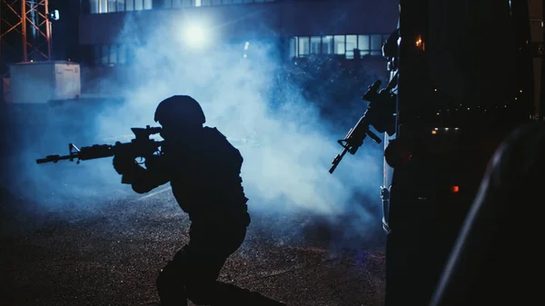 Silhouette eines maskierten Teams bewaffneter SWAT-Polizisten aus einem schwarzen Lieferwagen, der vor einem Bürogebäude geparkt ist. Soldaten mit Gewehren und Taschenlampen rennen über eine Straße voller Rauch. — Stockfoto