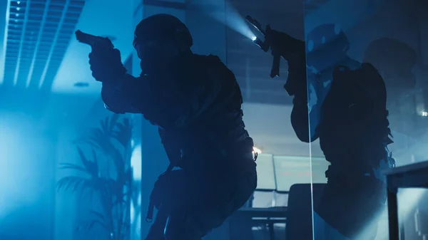 Команда озброєних поліцейських SWAT переміщується в залі будівлі темного прицілу з столами і комп'ютерами. Солдати з гвинтівками та ліхтарями "Сурвейл" та "Накриття". — стокове фото