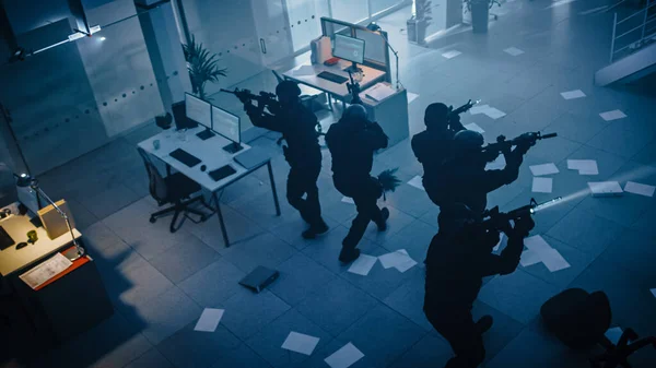 武装SWAT警察的蒙面小队用办公桌和计算机袭击了一幢黑暗扣押的办公楼。拿着步枪和闪光灯的士兵向前移动，掩护包围。以上摄象机. — 图库照片