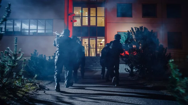 Equipe mascarada de policiais armados da SWAT correm em formação para um prédio de escritórios. Soldados com espingardas e lanternas funcionam em uma rua cheia de fumaça. — Fotografia de Stock