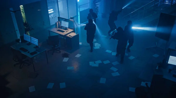 Maskeret trup af bevæbnede SWAT politifolk Storm en mørk beslaglagt kontorbygning med skriveborde og computere. Soldater med rifler og lommelygter bevæger sig fremad og dækker omgivelserne. Oversigtskamera. - Stock-foto