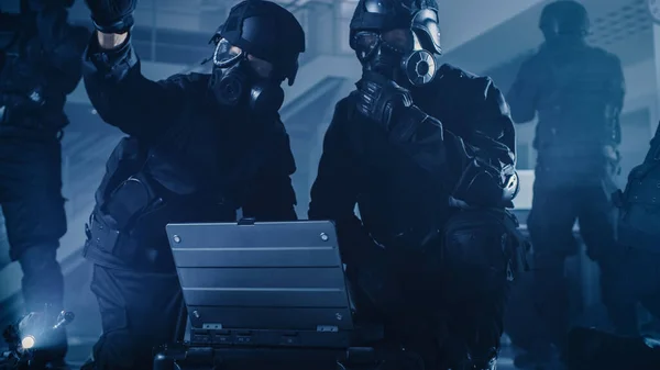 Maskierte Polizisten mit Gewehren stehen in einem dunklen Bürogebäude, in dem Schreibtische und Computer stehen. Soldat öffnet Laptop, um einen taktischen Angriff zu planen. — Stockfoto