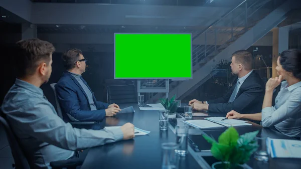 Sent i kveld i foretaksmøterommet: Styret for direktører, ledere og forretningsfolk Sitter ved forhandlingsbordet, snakker og bruker Green Mock-up Wall-TV for Video Conference Call. – stockfoto