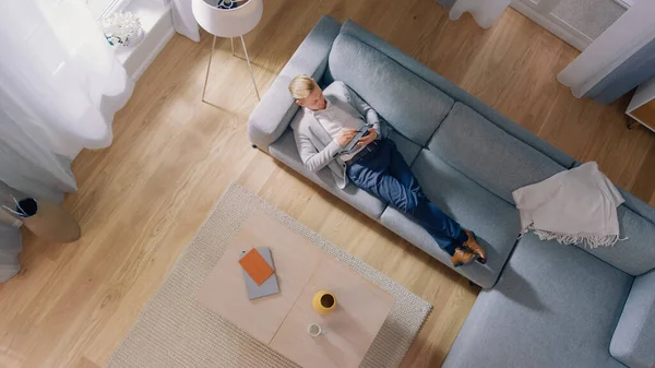 Le jeune homme est allongé sur un canapé, travaillant ou dessinant sur un ordinateur tablette. Salon confortable avec intérieur moderne, canapé gris et parquet. Vue du dessus Prise de vue caméra. — Photo