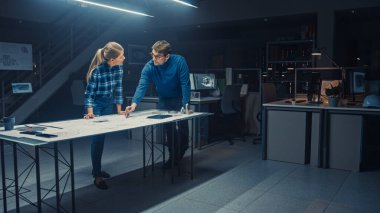 Karanlık Endüstriyel Tasarım Mühendislik Tesisi Erkek ve Kadın Mühendisler Konferans Masası Kullanarak Bir Planlar ve Teknik Çizimler Üzerinde Çalışıyor. Tesis Motor Kavramlarını Gösteren Bilgisayarlarla Doludur