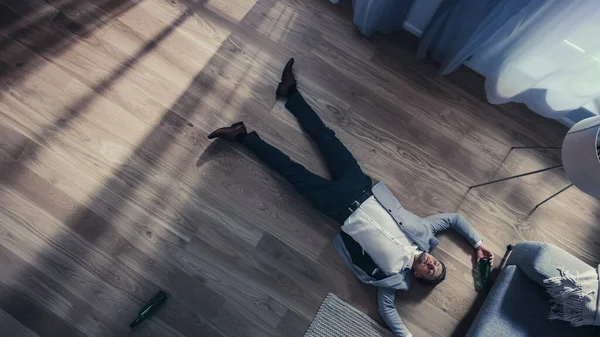 Poor Depressed Drunk Young Man is Crawling Towards a Sofa in an Apartment with Wooden Flooring. Hes Holding a Bottle of Beer and Tries to Drink the Leftovers. Dramatic Top View Camera Shot. — Stock Photo, Image