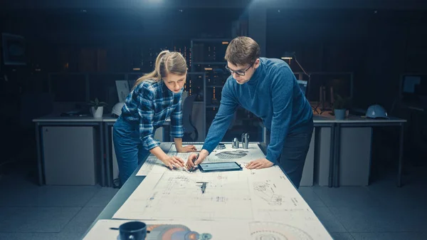 En la Instalación de Ingeniería de Diseño Industrial Oscuro Ingenieros Masculinos y Femeninos Hablan y Trabajan en un Plano Usando Mesa de Conferencias. Dibujos en el escritorio, borradores y componentes eléctricos del motor, piezas —  Fotos de Stock