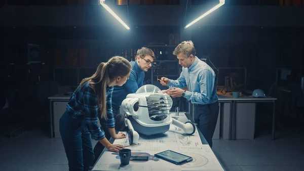 Tard dans la nuit dans l'installation d'ingénierie robotique Trois ingénieurs techniques parlent et travaillent sur un prototype de robot à roues. En arrière-plan Centre de recherche de haute technologie avec des écrans montrant le design industriel — Photo
