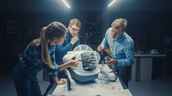 Tarde en la noche en las instalaciones de ingeniería de robótica Tres ingenieros técnicos hablan y trabajan en un prototipo de robot con ruedas. En el centro de investigación de alta tecnología de fondo con pantallas que muestran diseño industrial — Foto de Stock