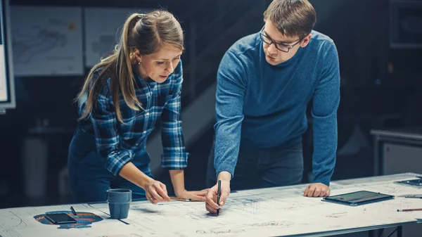 I Dark Industrial Design Engineering Facility Man och Kvinnliga Ingenjörer Prata och Arbeta på en ritning Använda Digital Tablet och Konferensbord. På skrivbordsritningar och motorkomponenter — Stockfoto