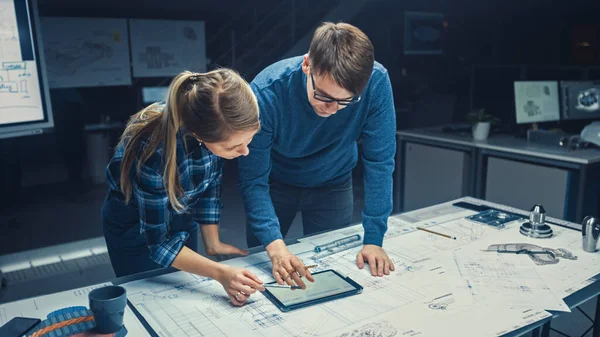 In der Dark Industrial Design Engineering Facility sprechen und arbeiten männliche und weibliche Ingenieure mit digitalem Tablet und Konferenztisch an einem Entwurf. Auf den Desktop-Zeichnungen und Motorkomponenten — Stockfoto