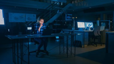 Masasında oturan güzel bayan mühendis bir dizüstü bilgisayarla çalışıyor. Bir Masanın Üzerinde Yatan Planlar. Karanlık Endüstriyel Tasarım Mühendislik Tesisinde.